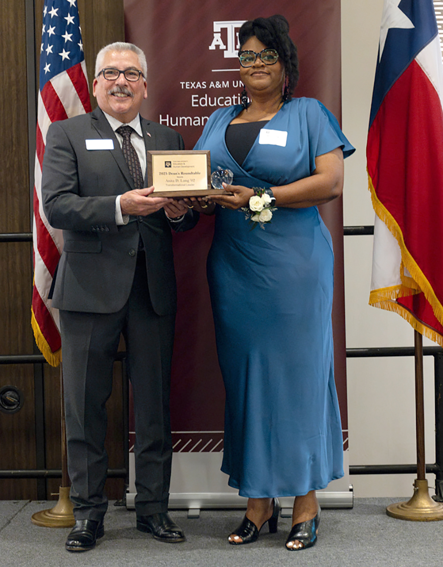 Two people stand side by side on a stage, smiling and holding a plaque together. On the left is a man in a dark gray suit with a white shirt, patterned tie, glasses, and gray hair and mustache. On the right is a woman in a teal blue satin dress with a white corsage on her wrist, glasses, and styled hair. Behind them is a Texas A&M University banner that reads “Texas A&M University Education and Human Development,” flanked by the U.S. and Texas flags. The plaque being presented reads “2023 Dean’s Roundtable Anita D. Long '92.”
